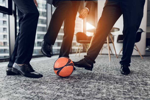 Close up. Three men in strict trousers and black shoes play an orange ball in football in the office - Zdjęcie, obraz