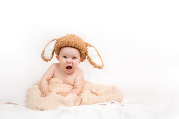A cute little baby wearing a bunny hat is sitting on the white bed, covered with a towel. White background. Copy space - Photo, Image