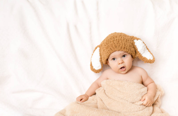 バニーの帽子をかぶってかわいい赤ちゃんはタオルで覆われて、白いベッドの上に座っています。白い背景。泣き叫ぶ子供。コピー スペース - 写真・画像
