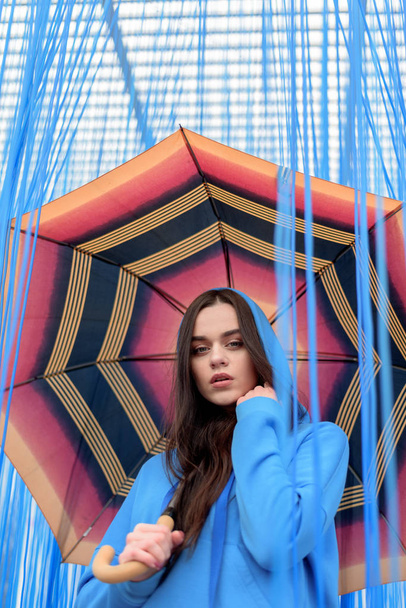 young beautiful woman with orange umbrella fashionably posing on the blue background. Rain concept - 写真・画像