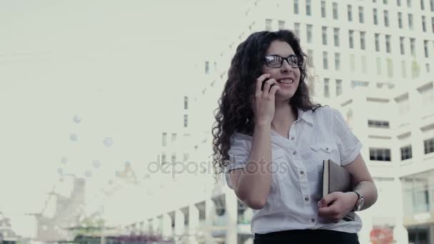 Jonge Aziatische meisje permanent op het stadsplein en praten over de telefoon. In handen model houden papieren notitieblok. Meisje met lang krullend haar gekleed in een wit overhemd, zwarte rok en glazen - Video