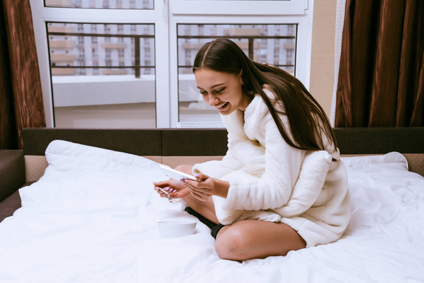 a laughing young girl in a white terry dressing gown is sitting on the bed and eating oatmeal porridge - Фото, изображение