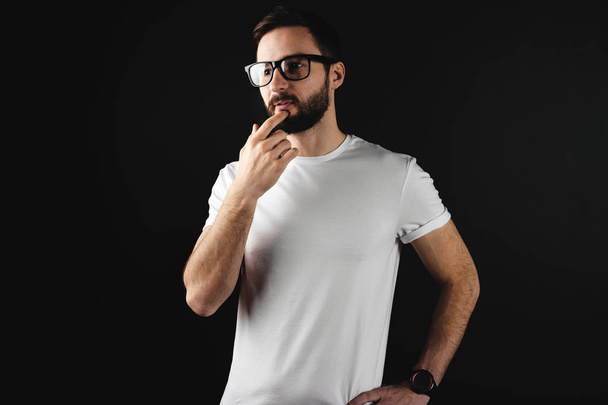 Pensive bearded guy in glasses wearing in white blank t-shirt on black background - Фото, изображение