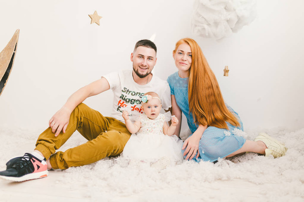 Un grupo de jóvenes, una familia de padre y madre de 30 años y una hija de un año, casas en un interior de luz se sientan en un suelo de madera, un fondo de una pared blanca y nieve artificial
. - Foto, imagen