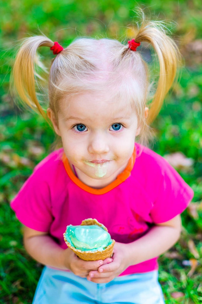 Забавная белая блондинка с голубыми глазами с двумя хвостами на голове, поедающая мороженое в вафельной чашке синего цвета, сидящей на зеленой траве. Все лицо грязное в растаявшем мороженом
 - Фото, изображение
