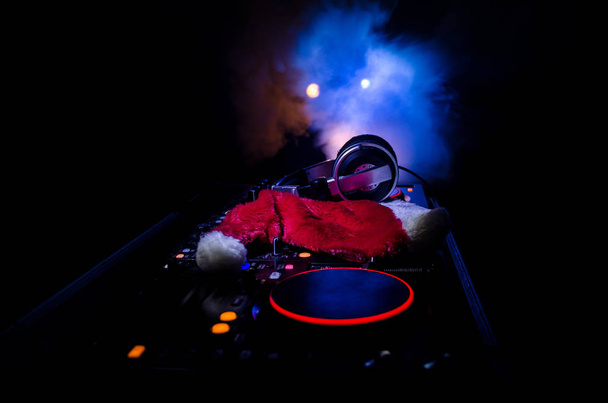 Mezclador de DJ con auriculares en el fondo oscuro club nocturno con árbol de Navidad Nochevieja. Vista de cerca de los elementos o símbolos de Año Nuevo (Santa Claus, muñeco de nieve, perro 2018, caja de regalo) en una mesa de Dj. tonificado
 - Foto, Imagen