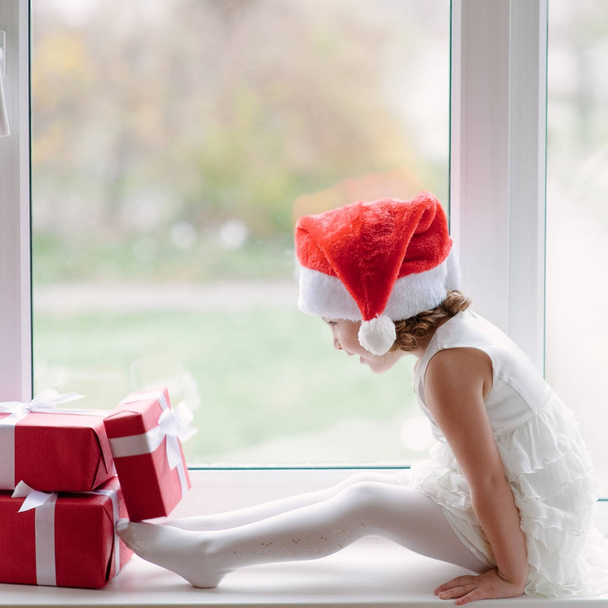 small girl in Santa hat sitting at the window with gifts - Fotoğraf, Görsel