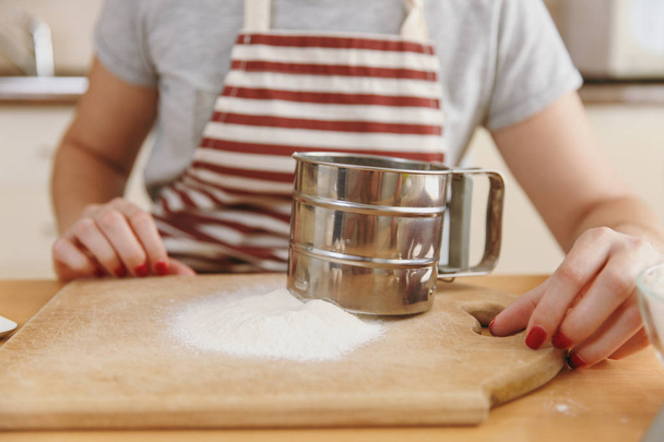 Close up shot of the young woman in apron with iron sieve and flour on the table in the kitchen. Cooking home. Prepare food. - Foto, Imagen