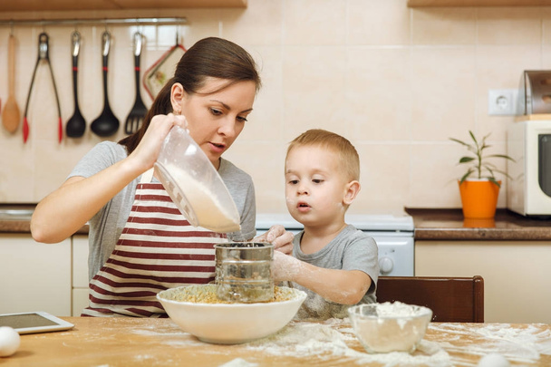 Jongen-jongetje helpt moeder om te koken kerst gember koekje in lichte keuken met tablet op tafel. Gelukkige familie moeder 30-35 jaar en kind 2-3 in weekend's ochtends thuis. Begrip van de relatie - Foto, afbeelding