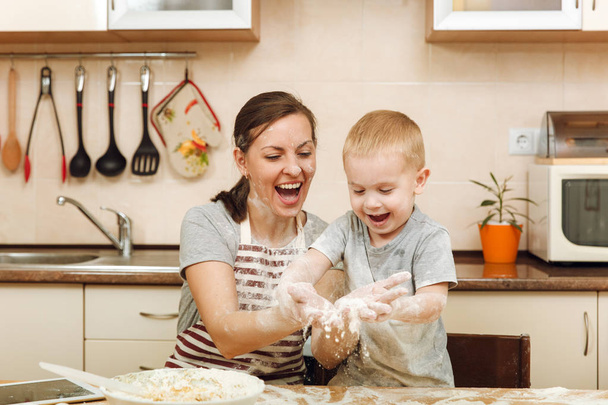 Kleiner Junge hilft Mutter, in heller Küche mit Tablette auf dem Tisch Ingwerkekse zu kochen. glückliche Familienmutter 30-35 Jahre und Kind 2-3 haben Spaß und werfen Mehl nach Hause. Beziehungskonzept - Foto, Bild