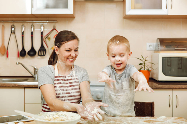 Little kid boy helps mother to cook Christmas ginger biscuit in light kitchen with tablet on the table. Happy family mom 30-35 years and child 2-3 have fun and throw flour home. Relationship concept - Foto, imagen