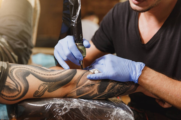 Close up of professional tattooer artist doing picture on hand of man by machine black ink from a jar. Tattoo art on body. Equipment for making tattoo art. Master makes tattooed in light studio. - Photo, Image