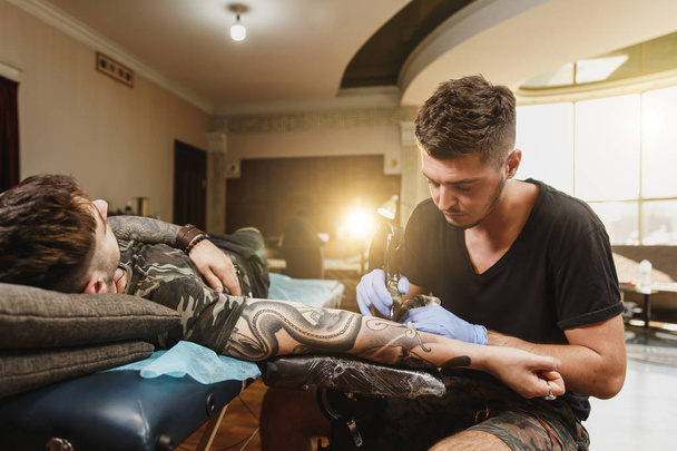 Tattooer profesjonalny artysta robi zdjęcie na dłoni człowieka przez maszyny czarny tusz od słoika. Sztuka tatuaż na ciele. Sprzęt do tatuażu sztuki. Master sprawia tatuażem w studio światła. - Zdjęcie, obraz