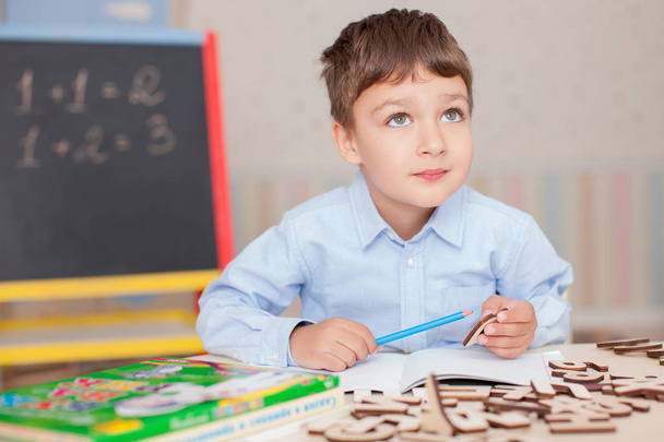 Cute little boy wearing blue shirt sitting at table and studying. Wooden letters, numbers, book copybooks and colorful pens and pencils on table. Blurred wall and blackboard background - Foto, Imagen