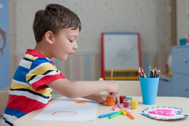 Happy child boy, wearing colorful t-shirt, drawing on white paper sheet, standing at table with colored paints and pencils - Photo, image