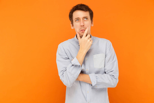 Hmm, i'm need thinking. Young adult man looking up, holding chin and musing. Indoor shot. Isolated on orange background - Photo, Image