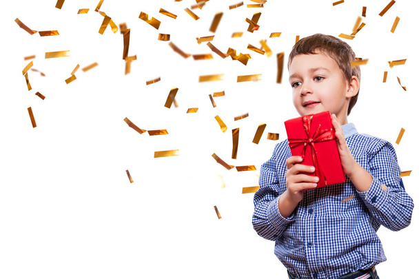 Cute child boy wearing jeans and blue shirt standing on white background with confetti and holding red gift in hands - Foto, immagini