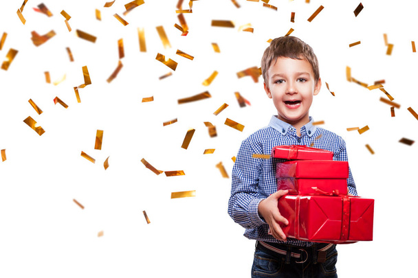Niño lindo con pantalones vaqueros y camisa azul de pie sobre fondo blanco con confeti y sosteniendo regalos rojos en las manos
 - Foto, Imagen