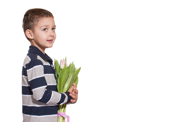 Studio shot of cute little boy wearing checkered shirt standing on white studio background with bouquet of pink tulips tied with pink ribbon in hands  - Fotó, kép