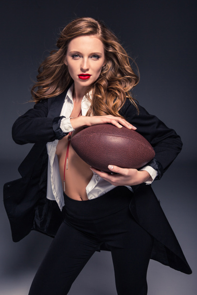 beautiful woman with long hair holding american football ball and looking at camera - Zdjęcie, obraz