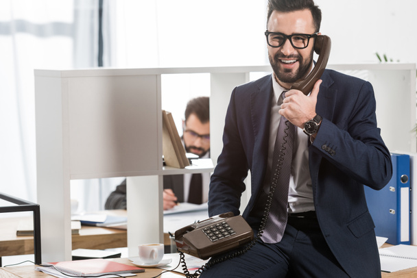 businessman talking by stationary telephone in office and sitting on table - Photo, Image