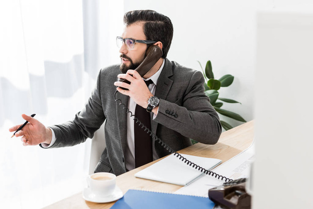 бизнесмен разговаривает по стационарному телефону в офисе и смотрит в сторону
 - Фото, изображение