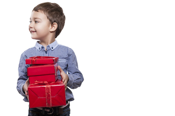 Studio shot of cute little boy wearing checkered shirt and dark jeans standing on white studio background with red gift boxes in hands  - Foto, Imagen