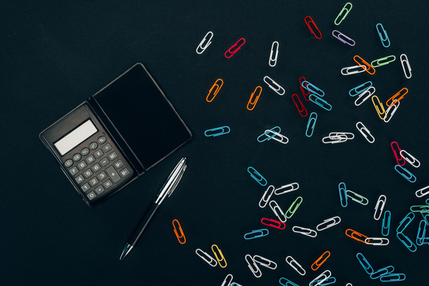 вид сверху калькулятора, перо и цветные скрепки на черном
  - Фото, изображение