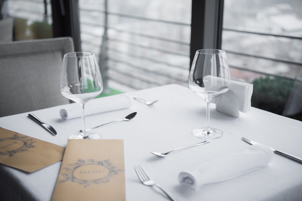 κλείνω πάνω θέα τοποθετημένα μαχαιροπήρουνα, άδειο wineglasses και μενού στον πίνακα με λευκό τραπεζομάντηλο σε εστιατόριο - Φωτογραφία, εικόνα