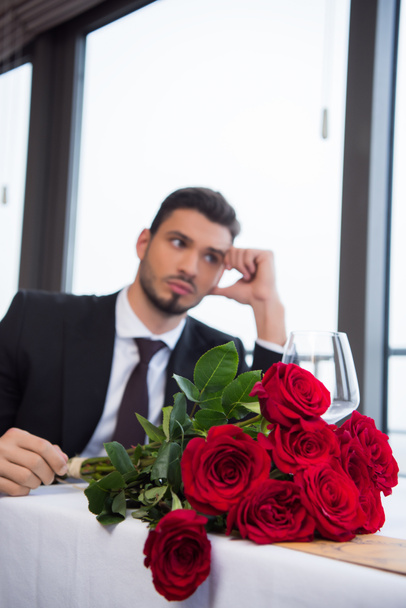 selective focus of man in suit with bouquet of red roses waiting for girlfriend in restaurant - Foto, afbeelding