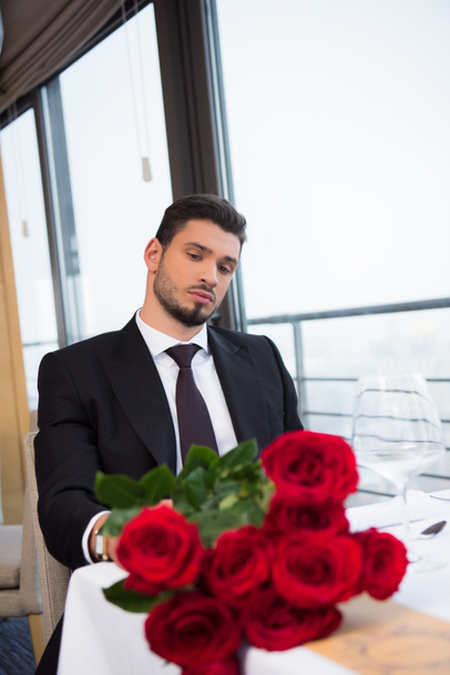 Πορτραίτο νεαρού κοστούμι με μπουκέτο από κόκκινα τριαντάφυλλα σε αναμονή για τη φίλη στο εστιατόριο - Φωτογραφία, εικόνα