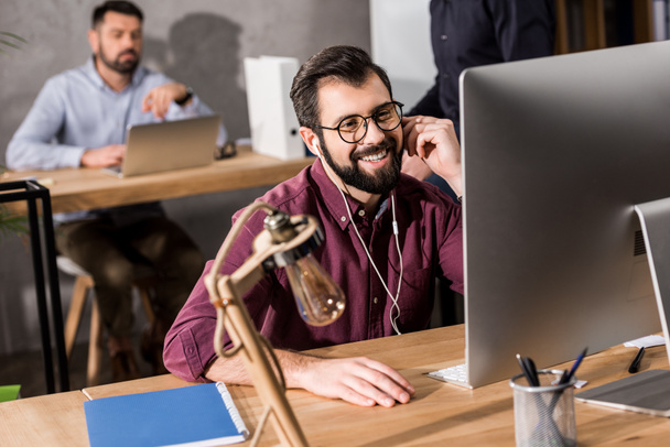улыбающийся бизнесмен, работающий за компьютером в офисе и слушающий музыку с наушниками
 - Фото, изображение