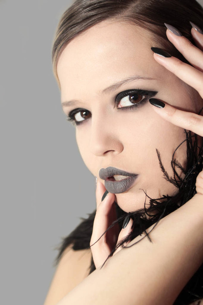 Maquiagem cinza preto fosco glamouroso, e manicuras em unhas nitidamente ovais com penas de um modelo em um fundo cinza
. - Foto, Imagem