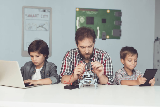 Two boys work on a tablet and a laptop, between them a man sits and constructs a robot - Photo, image