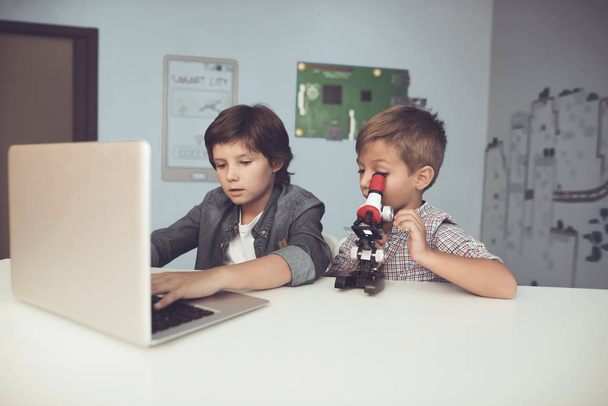 Two boys sitting at the table. One of them is sitting in front of a gray laptop. The second looks through a microscope - Fotoğraf, Görsel