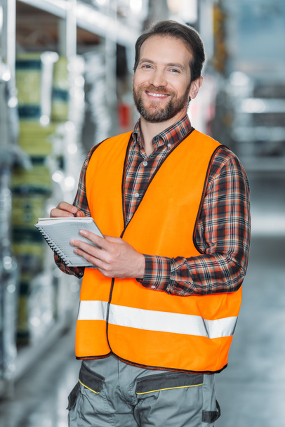 smiling worker in safety vest writing in notepad in storehouse - Photo, Image