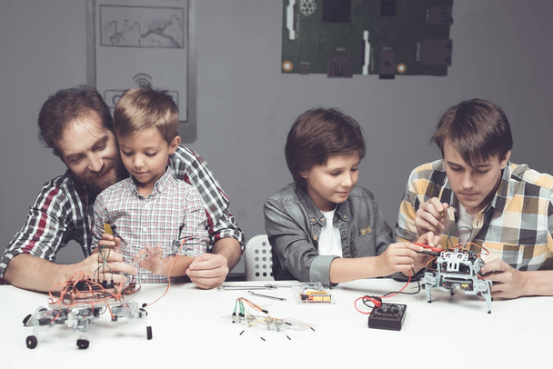 Two boys a teenager and an adult male are sitting in the workshop and constructing a robot - Fotoğraf, Görsel
