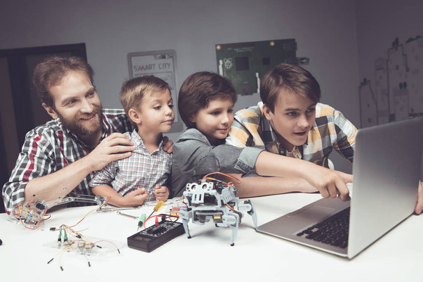 zwei Jungen, ein Teenager und ein erwachsener Mann sitzen in der Werkstatt und bauen einen Roboter - Foto, Bild