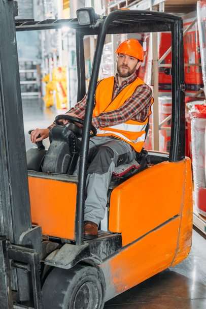 male worker in safety vest and helmet sitting in forklift machine in storage - Photo, Image