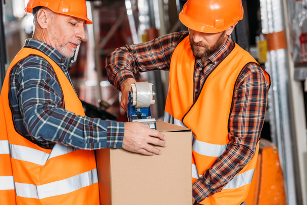mannelijke werknemers in Veiligheidsvesten en helmen verpakking kartonnen doos met plakband - Foto, afbeelding