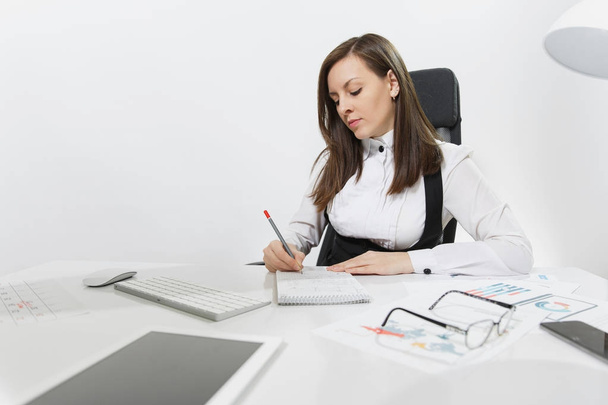Belle femme d'affaires sérieuse aux cheveux bruns en costume et lunettes assis au bureau, travaillant à l'ordinateur avec un moniteur moderne et des documents dans un bureau léger, écrivant dans un carnet sur fond blanc
 - Photo, image