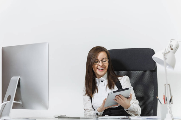 Bella donna d'affari dai capelli castani sorridente in abito e occhiali seduta alla scrivania, che lavora al computer con monitor moderno con documenti e tablet in ufficio leggero su sfondo bianco
 - Foto, immagini