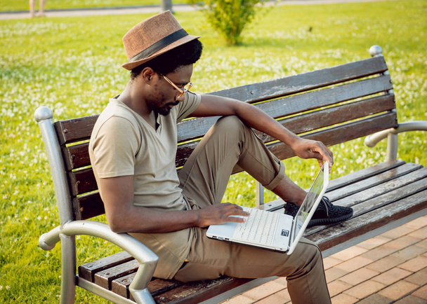 Young african man in hat and glasses with laptop in green city park - Φωτογραφία, εικόνα