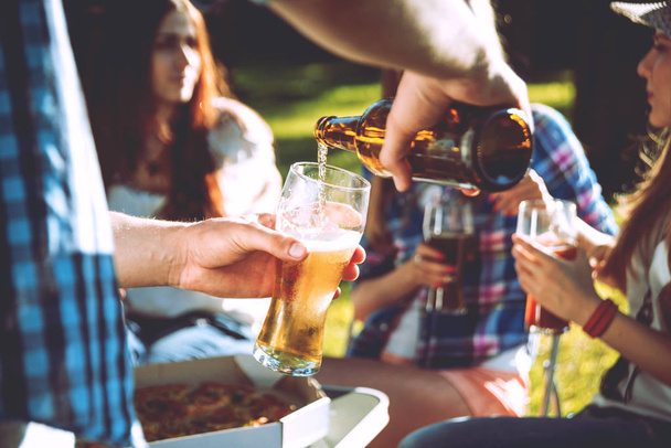 Cheerful caucasian friends on picnic in the park. Smiling and talking. Guy pours beer in a glass. - Photo, image