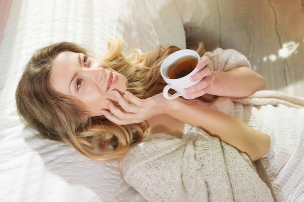 Closeup portrait of relaxing young woman enjoying coffee while sitting on floor near bed. Lazy morning in bed. - 写真・画像