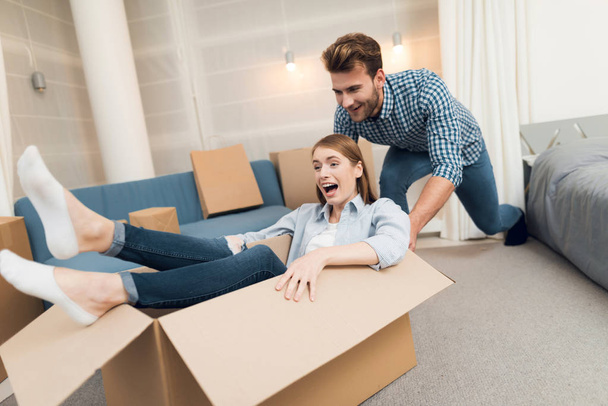 Young couple having fun while moving to new apartment. Moving newlyweds. Girl is sitting in a box. - Fotografie, Obrázek