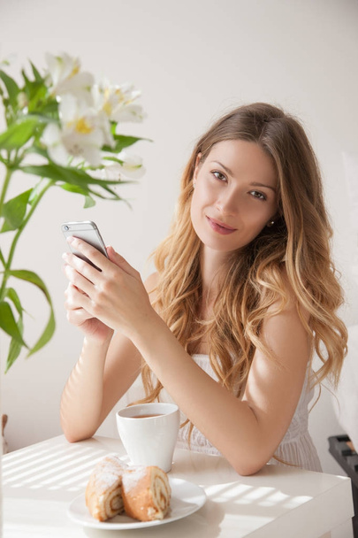 Beauty young woman using smartphone, young lady sitting at table with cup of tea and sliced apricots pie. Flowers in vase on table - Photo, Image