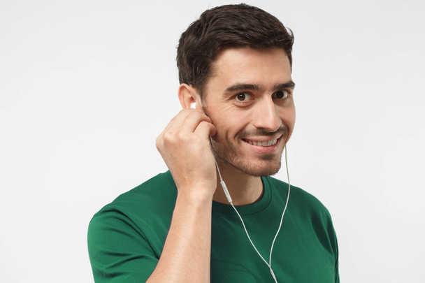 Handsome smiling man in green t-shirt listening to music with earphones, talking to friend via smartphone and headphones - Φωτογραφία, εικόνα