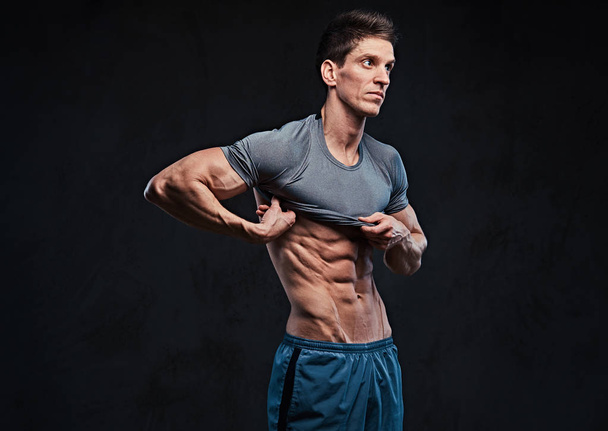Portrait studio d'ectomorphe muscle torse nu mâle enlève t-shirt et montre les muscles pectoraux sur fond gris foncé
. - Photo, image