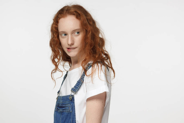 Indoor side portrait of young teenage girl with red curly hair isolated on white background standing in casual clothes looking behind her shoulder and biting her lip analyzing situation around - Foto, Imagen